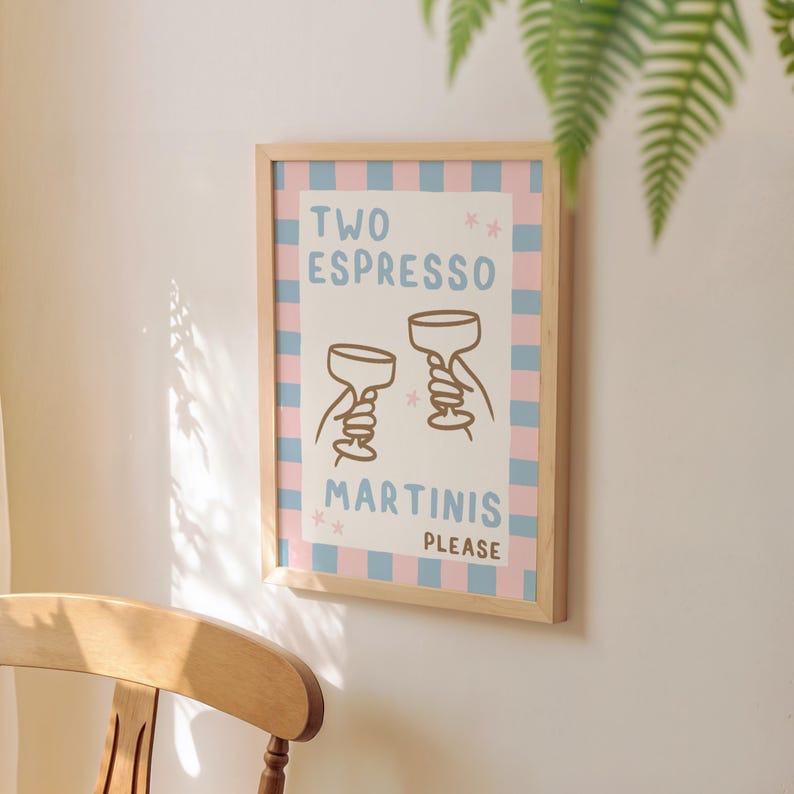 May include: A framed print with the text "TWO ESPRESSO MARTINIS PLEASE." The artwork features two cartoon martini glasses held by hands, set against a pink and blue checkered background. The frame is made of light-colored wood.