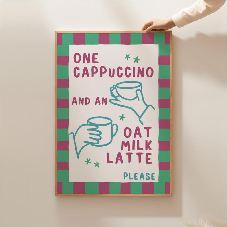 May include: A framed print with a green and pink striped border. The print features a white background with the text "One Cappuccino and an Oat Milk Latte Please" in teal blue lettering. The text is accompanied by two illustrations of hands holding cups of coffee.
