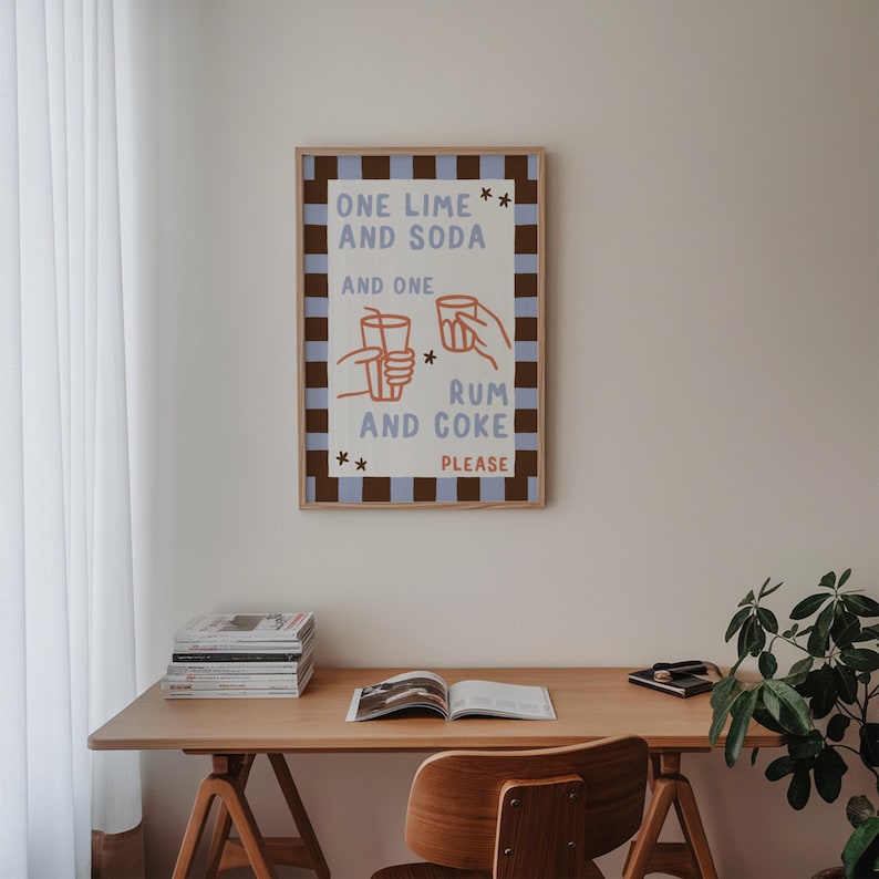May include: A framed art print with the text "ONE LIME AND SODA AND ONE RUM AND COKE PLEASE." The print has a light blue and brown checkered border and features illustrations of hands holding glasses. The print is hanging above a wooden desk.