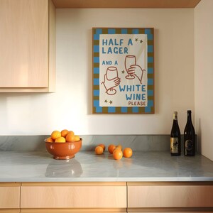 May include: A kitchen scene featuring a framed poster with the text "HALF A LAGER AND A WHITE WINE PLEASE." A terracotta bowl filled with oranges sits on a marble countertop, alongside several loose oranges and two wine bottles.