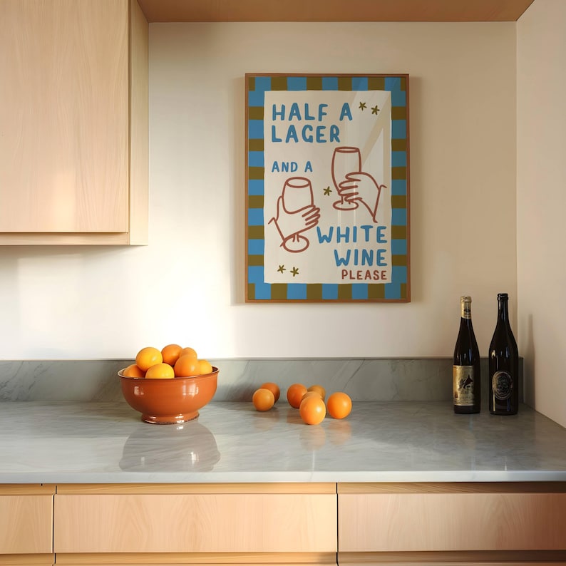 May include: A kitchen scene featuring a framed poster with the text "HALF A LAGER AND A WHITE WINE PLEASE." A terracotta bowl filled with oranges sits on a marble countertop, alongside several loose oranges and two wine bottles.