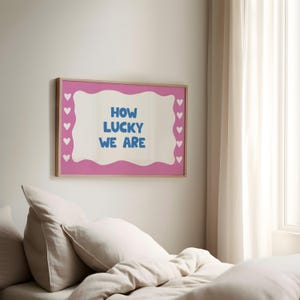 May include: A pink framed poster with a white background and blue text that reads "How lucky we are". The poster is surrounded by a border of white hearts.
