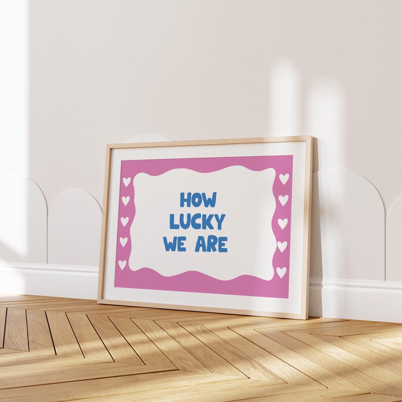 May include: A framed pink and white print with the text "HOW LUCKY WE ARE" in blue lettering. The print is surrounded by a white border and a white background.