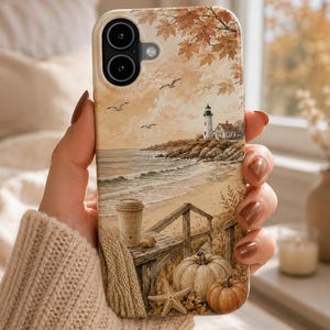 May include: A phone case featuring a coastal autumn scene with a lighthouse, a sandy beach, and seagulls. The foreground includes a wooden deck with a coffee cup labeled SALTY HAIR COZY VIBES, a starfish, and two pumpkins in warm, muted tones.