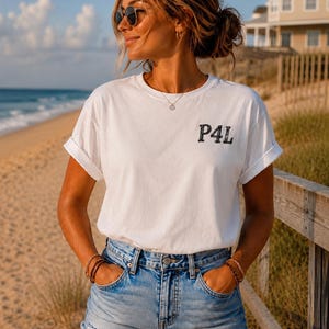 Pode incluir: Uma t-shirt branca de manga curta com gola redonda e mangas dobradas, apresentando o texto preto P4L impresso no lado esquerdo do peito, combinada com calções de ganga azul com bainha desfiada e detalhes desgastados na parte inferior.