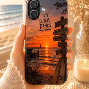 Könnte beinhalten: Eine Handyhülle mit einer Strand-Sonnenuntergangsszene und einem hölzernen Wegweiser mit der Aufschrift Outer Banks, Pogue Life, Kildare Island, The Cut, Popen und OBX. Ein Surfbrett lehnt am Pfosten und ein Leuchtturm ist in der Ferne zu sehen.