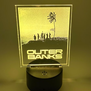 May include: A yellow illuminated acrylic sign with a silhouette of a group of people standing on a beach with a palm tree and the words "Outer Banks" below the image.