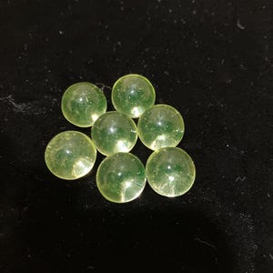 May include: Seven translucent, pale green marbles arranged on a black surface. The marbles are spherical and appear to be made of glass or a similar material. The lighting creates highlights and reflections on their surfaces.