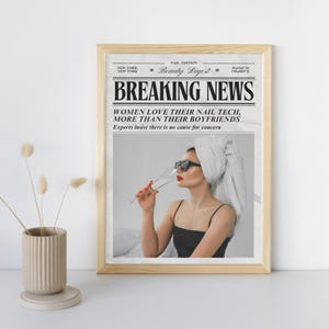 以下が含まれることがあります： 「BREAKING NEWS」という見出しの新聞風デザインの額入りプリント。サングラスとタオルを頭に巻いた女性がシャンパングラスを持っている画像です。「女性はボーイフレンドよりもネイル技術を愛している」という見出しです。