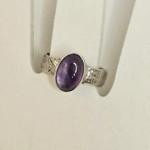May include: A silver ring featuring an oval, deep purple gemstone. The band has a textured design. The ring is displayed against a white background, highlighting the jewelry's details and craftsmanship.