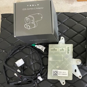 May include: A gray box with the text "TESLA CCS Combo 2 Adapter" on the front. The box contains a black cable with a white connector and a gray electronic component with a QR code and the text "P1595114-00-C SONTZ1176200757D".
