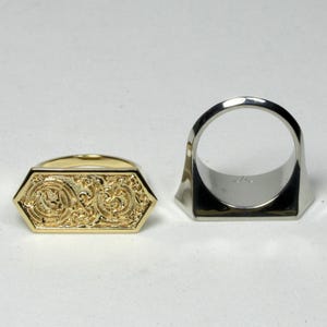 May include: Two rings: one gold-toned, hexagonal with ornate relief detailing, and one silver-toned, with a geometric design. The gold ring features a detailed pattern, while the silver ring has a simple, modern shape.