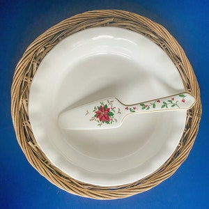 May include: A white pie plate with a scalloped edge sits on a woven wicker placemat. A white and gold cake server with a red poinsettia design rests on the plate.