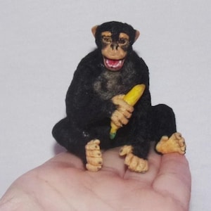 Op de afbeelding: Een klein beeldje van een zwarte chimpansee die zit en een gele banaan vasthoudt. De chimpansee heeft een lichtbruin gezicht, handen en voeten, met een open mond die tanden laat zien. Het figuurtje is getextureerd om op vacht te lijken.