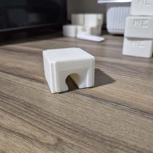 May include: A small, white, 3D-printed rectangular object with a curved opening on one side. The object is sitting on a wooden surface. Other similar objects are visible in the background.