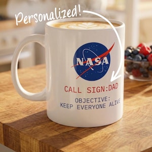 May include: A white ceramic mug filled with coffee featuring the blue and red NASA logo, the text CALL SIGN: DAD in red, and the text OBJECTIVE: KEEP EVERYONE ALIVE in blue. The word Personalized! is written in white script above the mug.