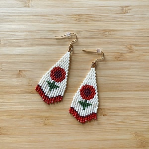 Könnte beinhalten: Ein Paar weiße Perlenohrringe mit roten und grünen Blumenmustern. Die Ohrringe sind tropfenförmig und hängen an goldenen Haken.