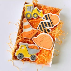 May include: A box of construction-themed biscuits. The biscuits are shaped like a yellow and grey excavator, a traffic cone, a dump truck, a hard hat, and a construction worker's vest. The biscuits are orange, yellow, grey, and white.