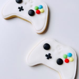 Può includere: Due biscotti bianchi di zucchero decorati per assomigliare a controller di videogiochi. Ogni biscotto ha pulsanti neri, un segno più nero e spolverate rotonde colorate.