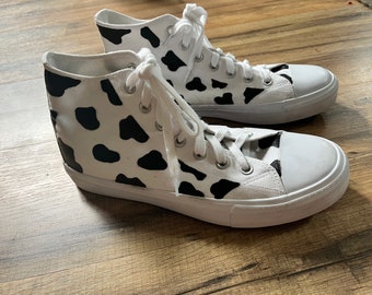 May include: A pair of white high-top canvas sneakers with a black and white cow print design.