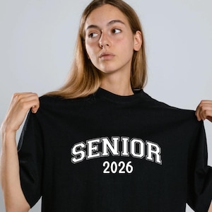 May include: Black t-shirt featuring the word "SENIOR" in white, curved text above the number "2026". The shirt has a crew neck design.