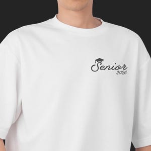 May include: White t-shirt with a black graduation cap and the word "Senior" and the year "2026" printed on the front. The shirt has short sleeves and a crew neck.