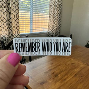 May include: A white sticker with the text "REMEMBER WHO YOU ARE" in black lettering. The sticker is being held by a hand with pink nail polish.