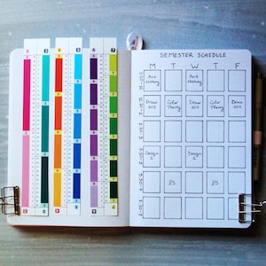 May include: An open notebook featuring a hand-drawn semester schedule grid with columns for Monday through Friday and rows for time slots. Three colorful vertical bookmarks with numbered sections are attached to the left page, and a pen is clipped to the right.