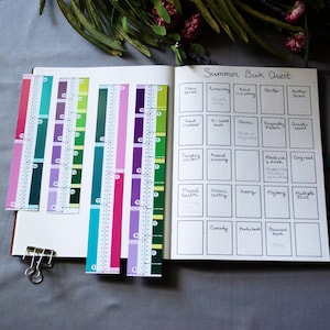 May include: An open notebook with a Summer Book Quest grid containing handwritten categories and titles. Beside the notebook are four colorful paper bookmarks with printed inch-based rulers, featuring shades of teal, purple, green, and pink.