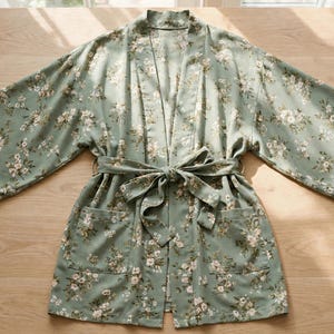 May include: A sage green kimono-style robe featuring a delicate white and brown floral pattern throughout. The garment includes wide three-quarter length sleeves, a matching fabric tie belt at the waist, and two front patch pockets.