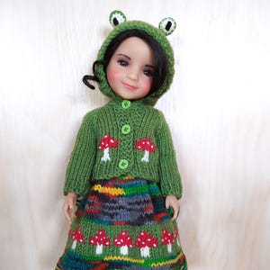 Puede incluir: Una muñeca vestida con un conjunto de punto verde con capucha de rana y detalles de setas. El conjunto incluye una rebeca con botones y una falda con diseños de setas rojas y blancas. La muñeca lleva botas plateadas.