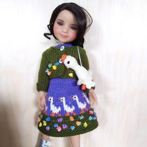 Puede incluir: Muñeca con un suéter verde, una falda morada con gansos blancos y botas plateadas. La muñeca tiene cabello negro y lleva un bolso con forma de ganso blanco. La falda y el suéter están adornados con detalles florales coloridos.