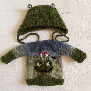 Könnte beinhalten: Gestricktes Baby-Outfit im Frosch-Design, bestehend aus einem grünen Hut mit Augendetails und einem passenden Pullover. Der Pullover zeigt ein Froschgesicht und Pilz-Akzente. Für Säuglinge.
