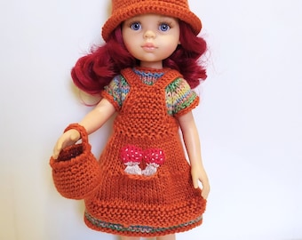 May include: A doll wearing a knitted orange dress with a mushroom design, a matching orange hat with white polka dots, and orange shoes. The doll has red hair and blue eyes.