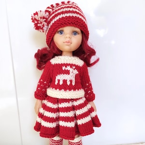 Può includere: Una bambola che indossa un completo lavorato a maglia rosso e bianco, tra cui un cappello con pompon, un maglione con un motivo a renna, una gonna e scaldamuscoli. La bambola ha i capelli rossi e scarpe verdi.