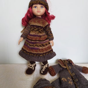 May include: A doll wearing a brown and purple striped knitted dress with a brown knitted hat. The doll has red hair and blue eyes. The doll is also wearing brown boots. The doll is holding a brown knitted jacket with buttons.