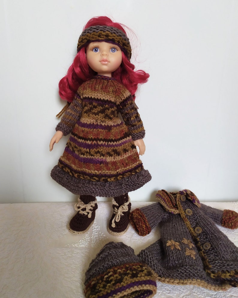 May include: A doll wearing a brown and purple striped knitted dress with a brown knitted hat. The doll has red hair and blue eyes. The doll is also wearing brown boots. The doll is holding a brown knitted jacket with buttons.