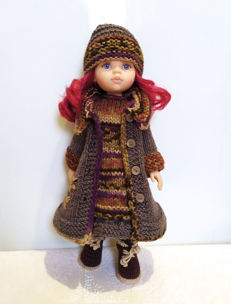 May include: A doll wearing a brown knitted coat with a striped pattern, a matching hat, and brown boots. The doll has pink hair and blue eyes.