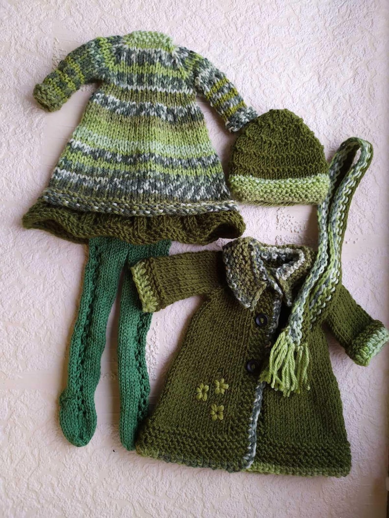May include: A set of knitted clothing for a doll, including a green and white striped dress, a green coat with a button closure, a green hat, and green tights.