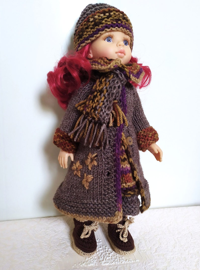May include: A doll wearing a brown knitted coat with a purple and brown scarf, a brown knitted hat with a purple and brown pattern, and brown knitted boots.