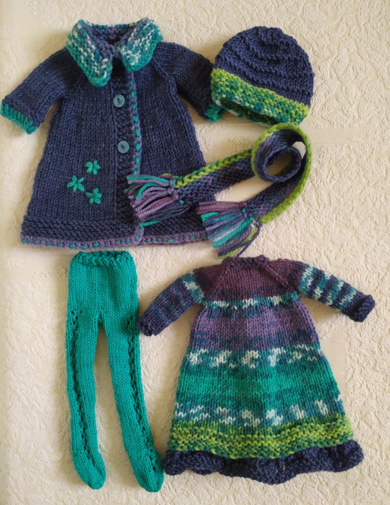 May include: A set of knitted doll clothes in shades of blue, green, and purple. The set includes a coat, dress, leggings, hat, and scarf.