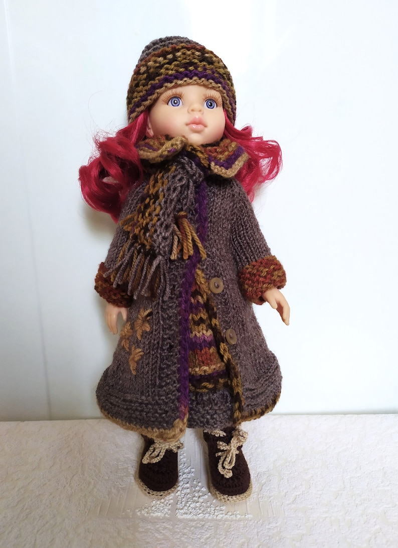 May include: A doll wearing a brown knitted coat with a colorful scarf, a brown knitted hat with purple and green stripes, and brown knitted shoes.