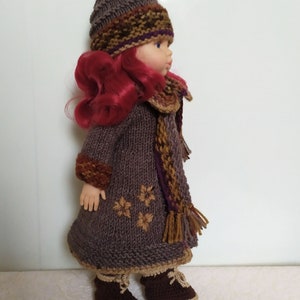 May include: A brown knitted doll outfit with a matching hat and scarf. The outfit features a brown knitted coat with a brown and gold scarf and a brown knitted hat with a brown, gold, and purple trim. The doll is wearing brown boots.