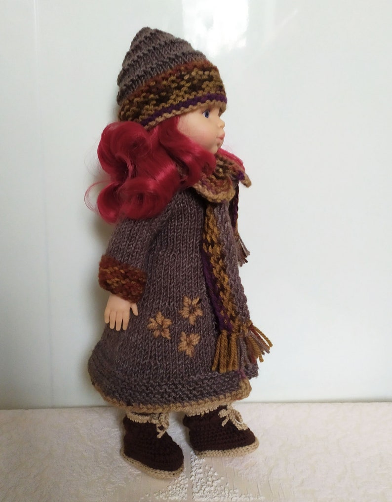 May include: A brown knitted doll outfit with a matching hat and scarf. The outfit features a brown knitted coat with a brown and gold scarf and a brown knitted hat with a brown, gold, and purple trim. The doll is wearing brown boots.