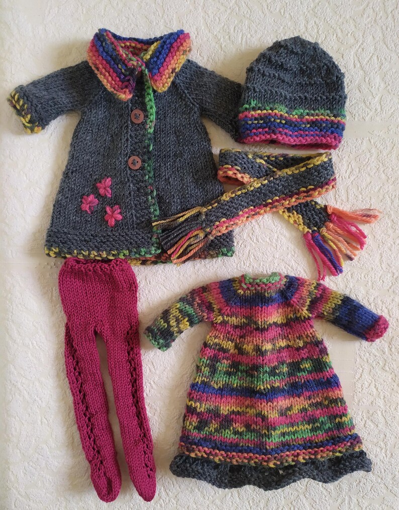 May include: A set of knitted doll clothes in grey, pink, and multi-colored stripes. The set includes a coat, hat, scarf, dress, and leggings.