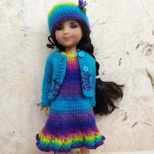 May include: A doll wearing a hand-knitted outfit. The outfit includes a blue, purple, and yellow dress, a matching blue jacket, and a hat with a purple flower. The doll has long black hair and is wearing a colorful outfit.