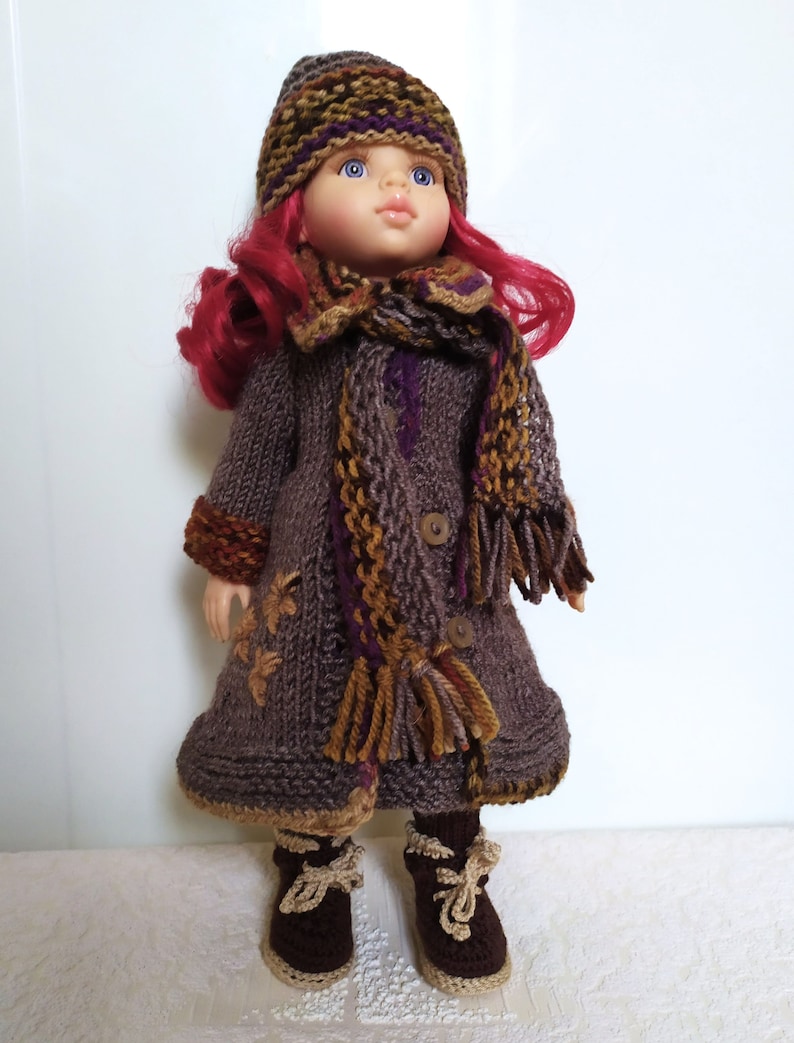 May include: A doll wearing a brown knitted coat with a purple and brown scarf, a brown knitted hat with purple and brown stripes, and brown knitted boots with tan laces.