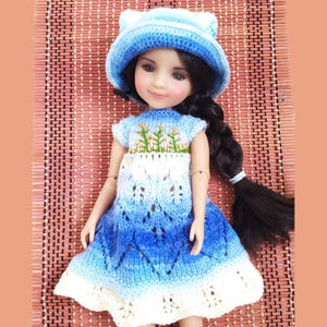 May include: A doll dressed in a blue and white knitted dress and a matching hat with cat ears. The dress has a floral design on the bodice and a gradient effect. The doll has long black hair in a braid.