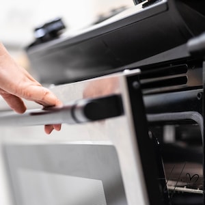 May include: Close-up of a stainless steel oven with a black handle. The oven door is partially open, revealing the interior. A hand is reaching for the handle. The oven is in a kitchen setting.