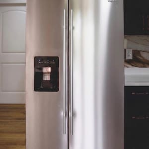 May include: Stainless steel Whirlpool refrigerator with a side-by-side door design. Features include a water and ice dispenser on the left door, and long, vertical handles. The refrigerator is in a kitchen setting with a white door and wood flooring visible.